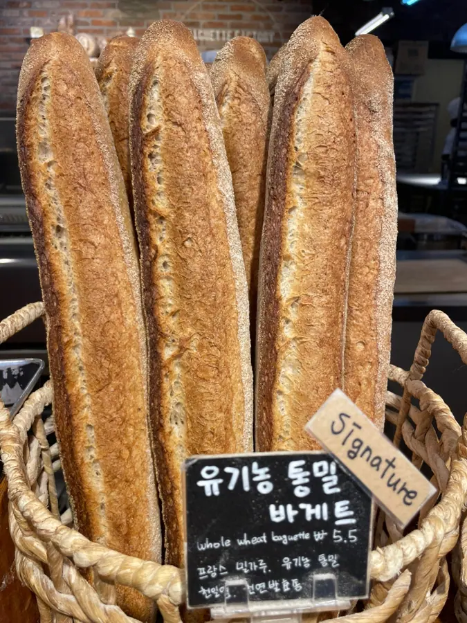 바게트제작소의 바구니에 유기농 통밀 바게트가 세워져 있고 앞에 검은 칠판이 놓여 있다. 칠판에 유기농 통밀 바게트라고 적혀 있다.