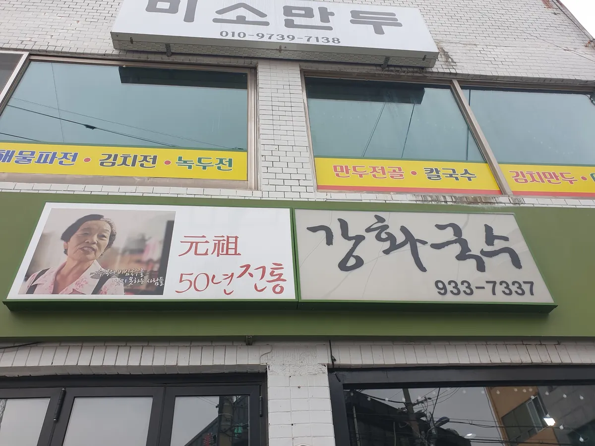 강화국수 간판. 손글씨로 강화국수라고 쓰고 아래 933-7337이 붙어 있다. 옆 간판에 元祖 50년전통이라고 적혀 있다.
