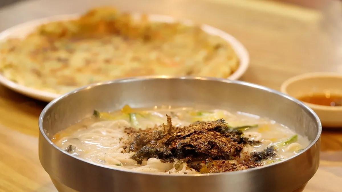 백령도 굴메밀칼국수 한 상. 스테인리스 대접에 담긴 메밀칼국수 위에 김가루와 들깻가루가 얹혀 있고, 뒤에 노릇하게 부친 전 한 접시와 간장 종지가 놓여 있다.