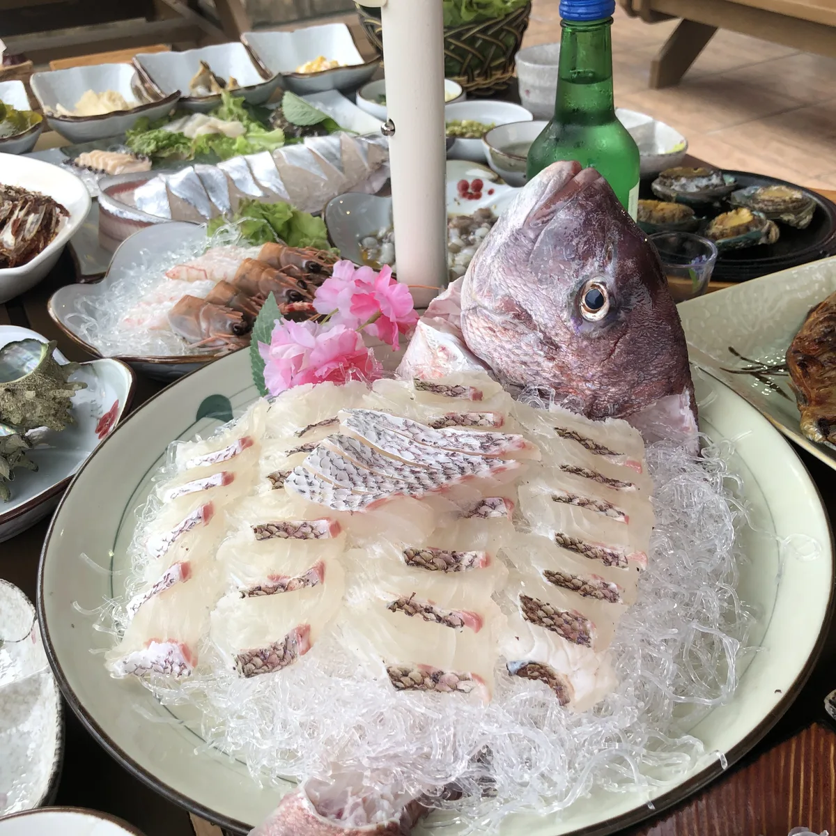 상 전체. 머리를 세워 담은 큰 생선회 접시에 흰 살 회가 겹겹이 깔렸고 뒤로 밑반찬과 소주병 하나가 놓여 있다.