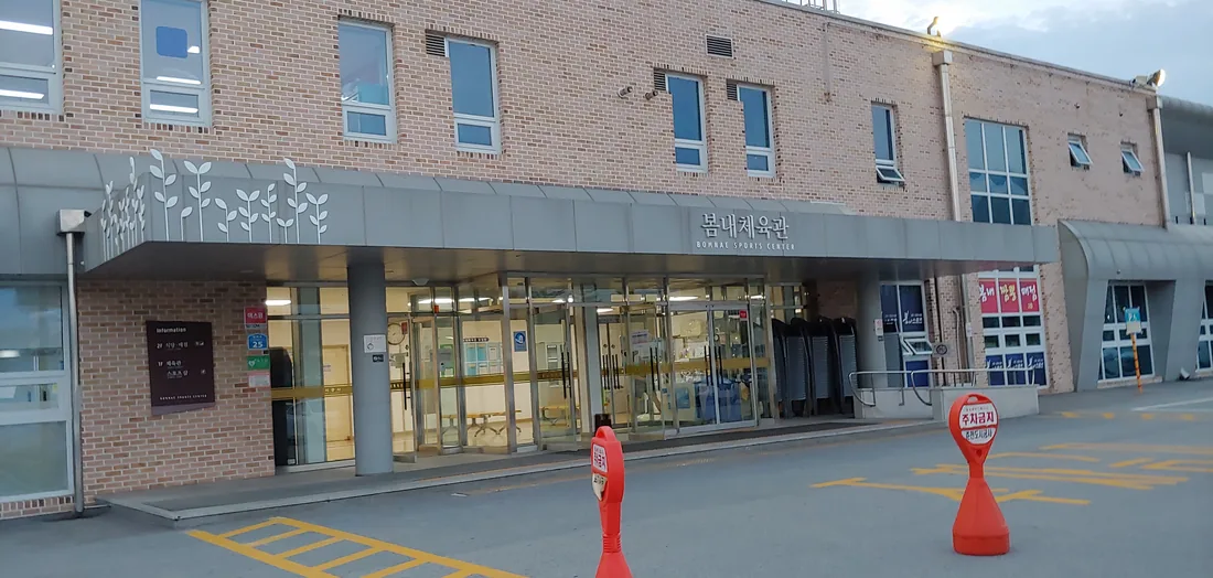 봄내체육관 정문. 벽돌 건물 처마에 봄내체육관과 BOMNAE SPORTS CENTER라고 적혀 있고, 왼쪽 안내판에 1층 체육관과 스포츠샵, 2층 식당과 매점이라고 표시되어 있다.