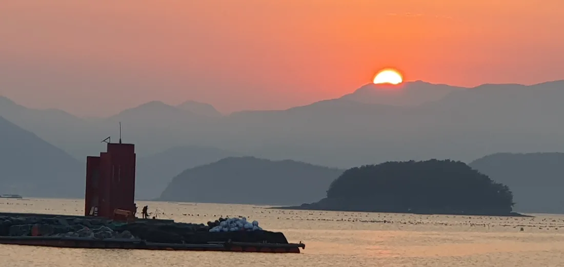 고성 앞바다에 해가 산등성이로 내려앉는 모습. 앞쪽 방파제에 붉은 등대가 서 있고 바다에는 양식장 부표가 줄지어 떠 있다.
