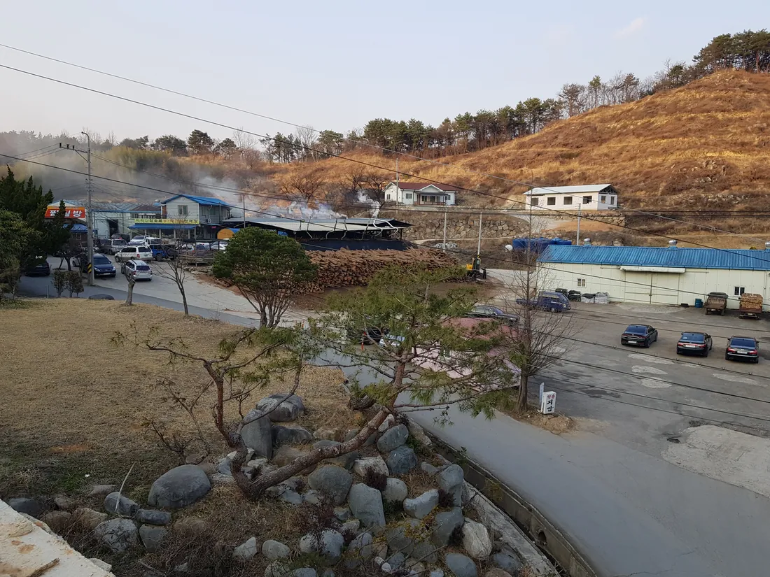산자락 아래 숯가마에서 연기가 오르고 옆에 통나무가 산더미로 쌓여 있다. 같은 마당의 고깃집은 생고기(삼겹살·목살) 11,000원, 양념갈비 12,000원, 생갈비 13,000원이다.