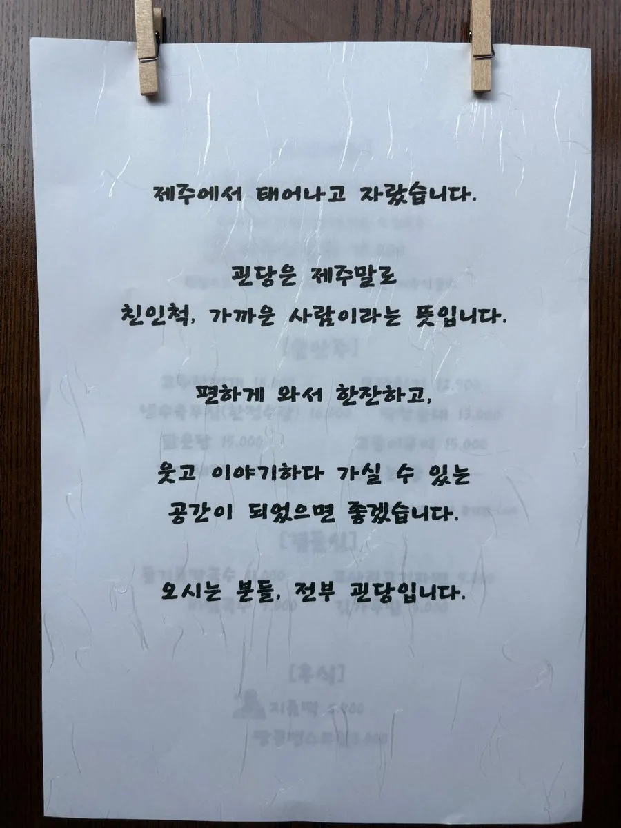 괸당 신당본점 벽에 집게로 매단 손글씨 안내문. 제주에서 나고 자랐다는 이야기와 괸당이 제주말로 친인척, 가까운 사람이라는 뜻이라는 설명이 적혀 있다.