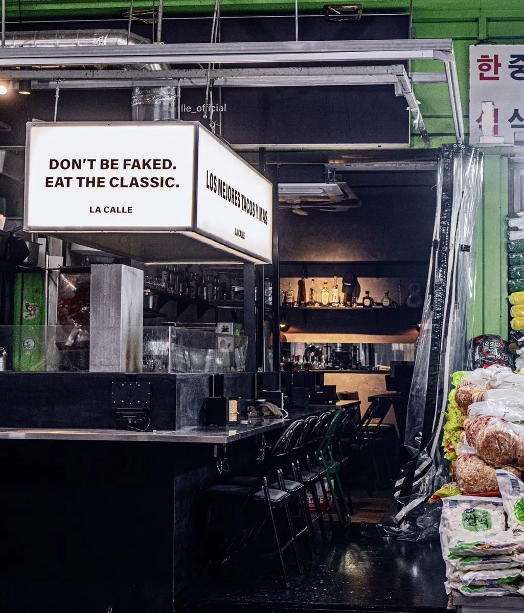 라까예 가게 앞. 「DON'T BE FAKED. EAT THE CLASSIC. LA CALLE」와 「LOS MEJORES TACOS Y MAS」라고 적힌 조명 간판 두 개가 걸려 있고 오른쪽에 시장 좌판 자루가 그대로 붙어 찍혔다.