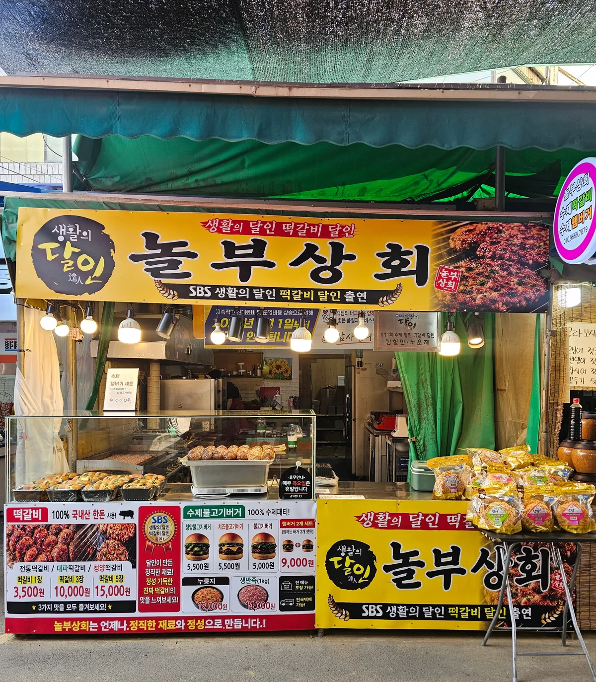 놀부상회 가게 앞. 시장 천막 아래 노란 간판에 「놀부상회」와 「SBS 생활의 달인 떡갈비 달인 출연」이 적혀 있고, 좌판 앞 배너에 떡갈비 3,500원 10,000원 15,000원과 버거 세트 9,000원이 붙어 있다.
