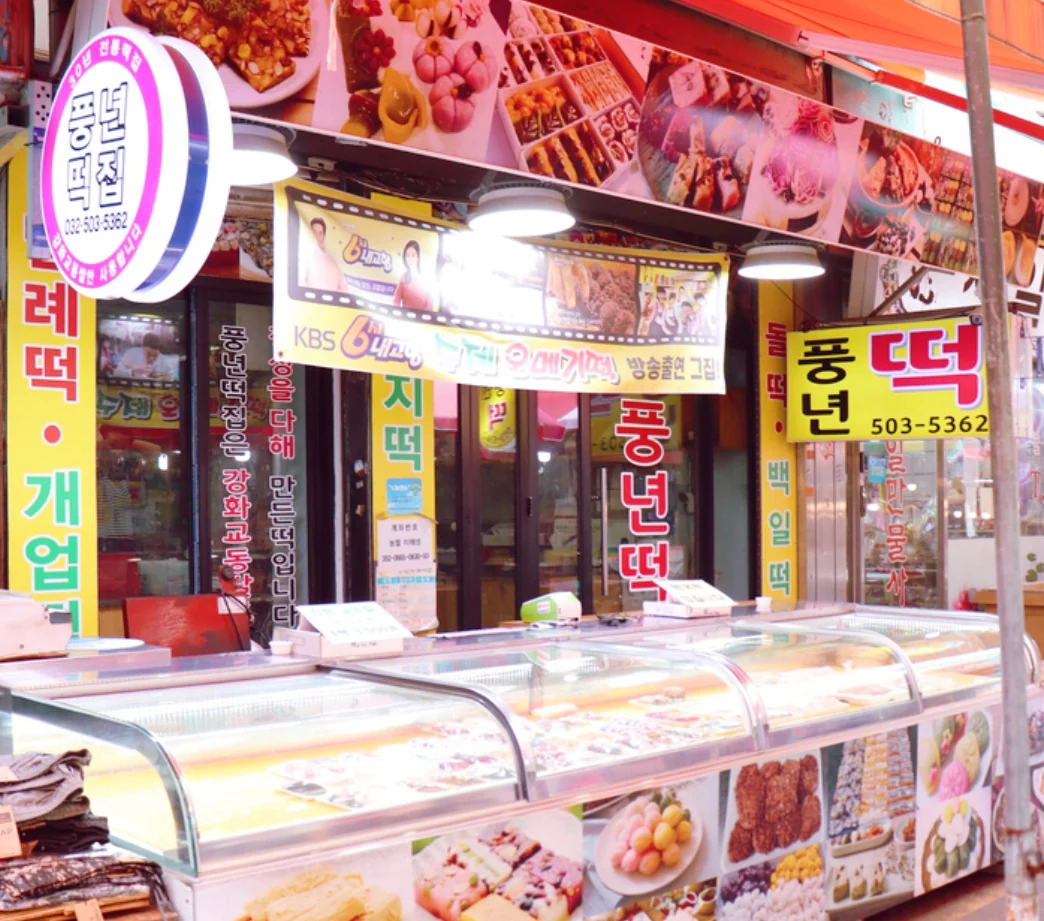 풍년떡집 가게 앞. 노란 「풍년떡」 간판이 걸려 있고 그 아래 진열장에 떡이 놓여 있다. 간판에 적힌 번호는 503-5362.