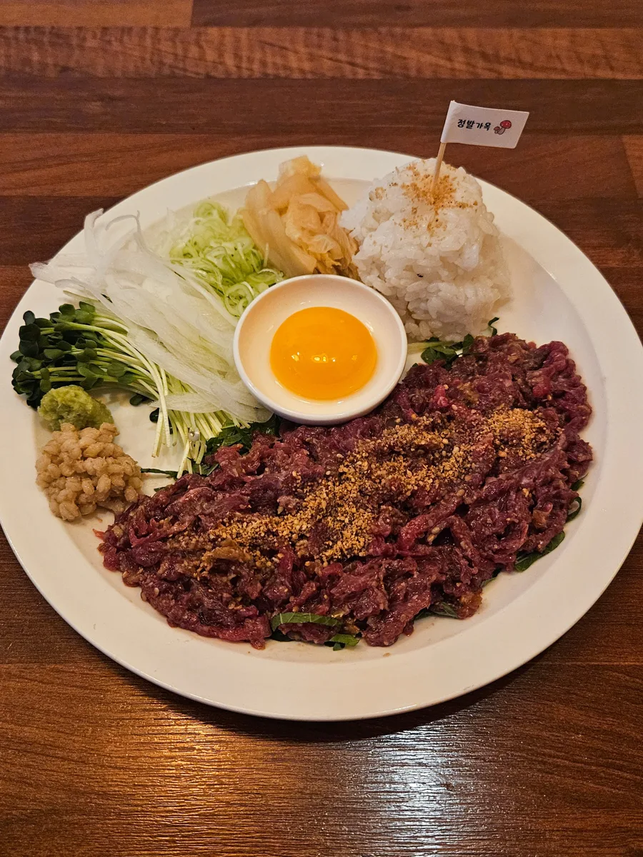정발가옥이 네이버 플레이스에 올린 한우 육회 플래터 사진. 접시 가운데 종지에 노른자를 담고 그 둘레로 육회를 펼쳤으며 백김치와 채 썬 배, 무순, 잣, 고추냉이를 곁들였고 밥 위에 정발가옥이라고 적힌 작은 깃발이 꽂혀 있다.