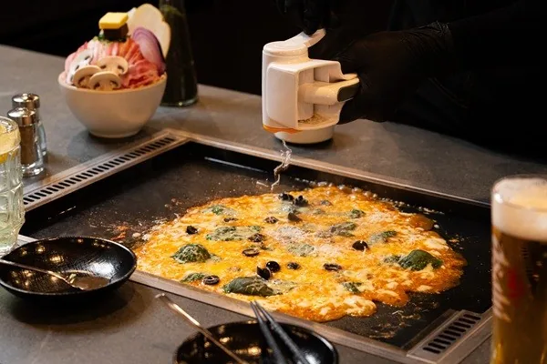 몬몬 몬자야끼 테이블 철판에 몬자야키가 넓게 펴져 익고 있고 그 위로 검은 장갑을 낀 손이 통을 들어 소스를 뿌린다.