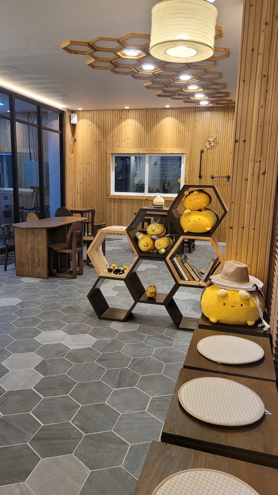 cafe벌통 안쪽 나무 방. 육각 선반에 노란 꿀벌 인형이 앉아 있고 책이 꽂힌 칸도 있으며 오른쪽에 좌식 방석이 놓인 낮은 상이 이어진다.