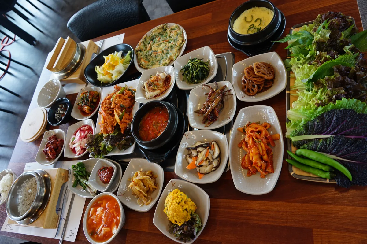 나무 상 위에 흰 접시 열댓 개가 빼곡히 놓여 있다. 연근조림, 가지, 오징어초무침, 계란말이, 표고버섯볶음, 부추전, 뚝배기 계란찜이 보이고 오른쪽에 적상추와 깻잎, 풋고추가 쟁반에 담겨 있다.
