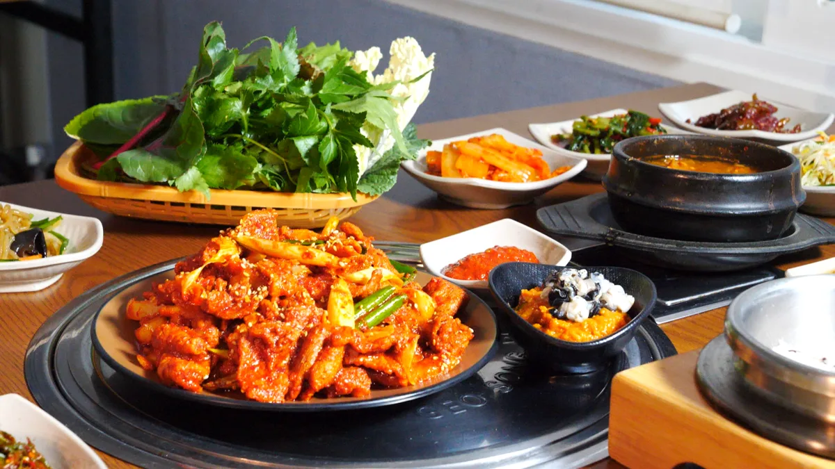 검은 무쇠판 위에 매운 양념 제육볶음이 수북이 담겨 있고, 뒤쪽 광주리에 깻잎과 상추 같은 쌈채소가 쌓여 있다. 상 가장자리로 나물과 무생채, 된장찌개 뚝배기가 둘러 있다.