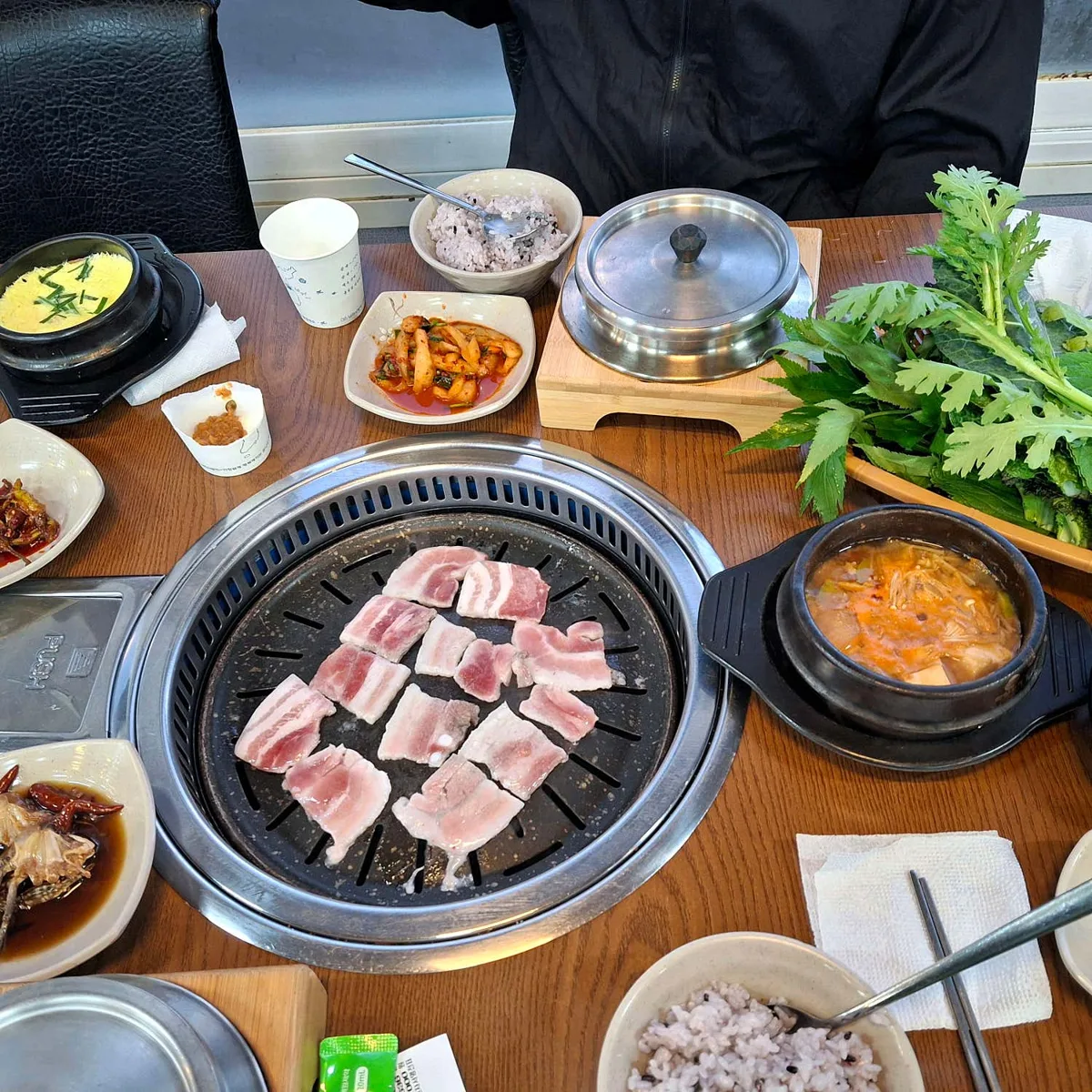 가운데 둥근 불판에 얇게 썬 냉동 삼겹살 열 점이 올라가 있다. 옆으로 뚝배기 계란찜과 된장찌개, 흑미 섞인 잡곡밥, 쑥갓과 케일이 담긴 쌈채소 쟁반이 놓여 있다.