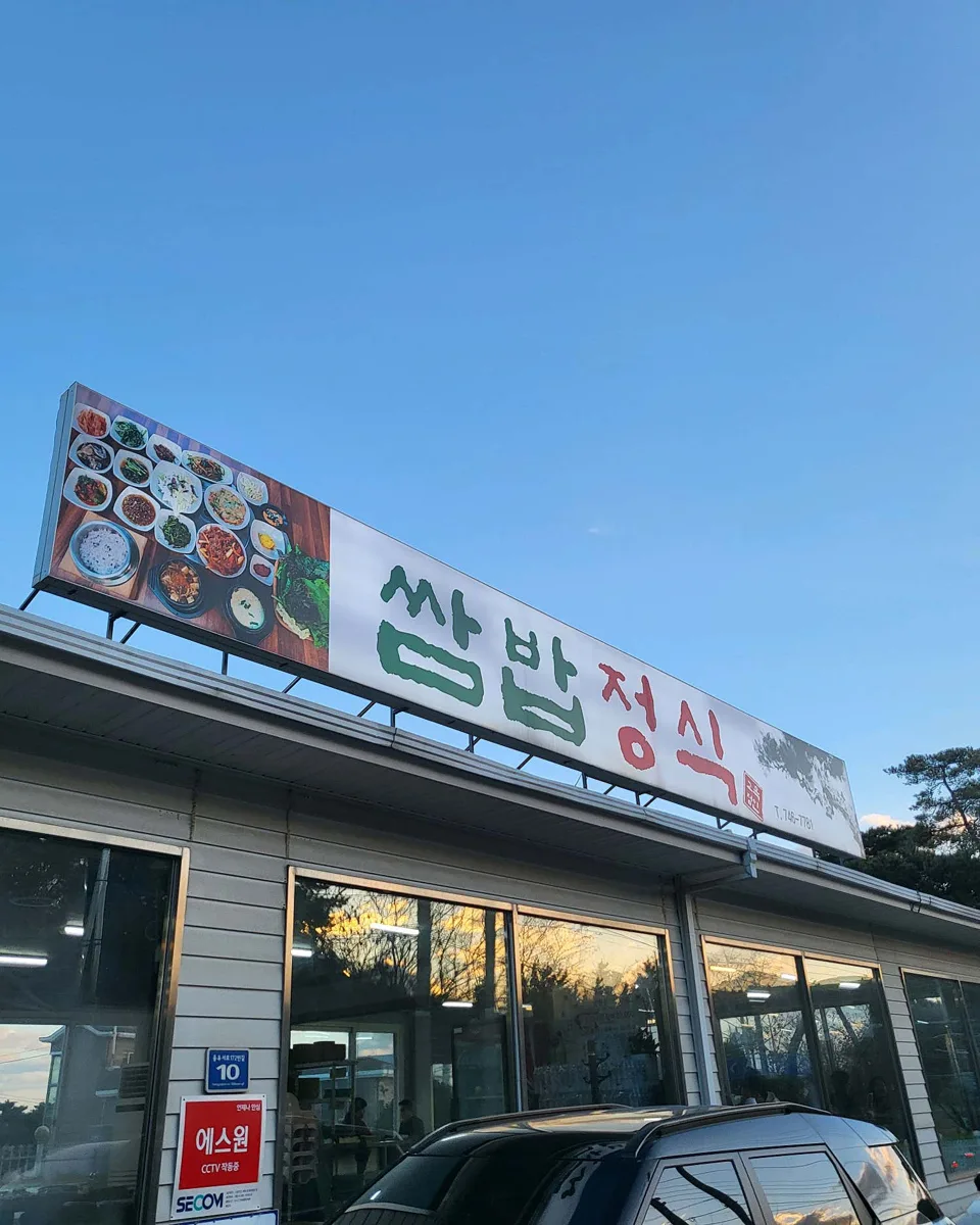 단층 건물 지붕 위에 쌈밥정식이라고 크게 쓴 간판이 올라가 있다. 간판 왼쪽에는 상차림 사진이 붙어 있고 건물 앞으로 차가 세워져 있다.